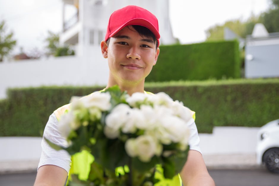 A delivery person wearing a red cap and a high-visibility vest is smiling and extending their arm towards the camera, holding a bouquet of fresh white flowers with green foliage. The bouquet appears t