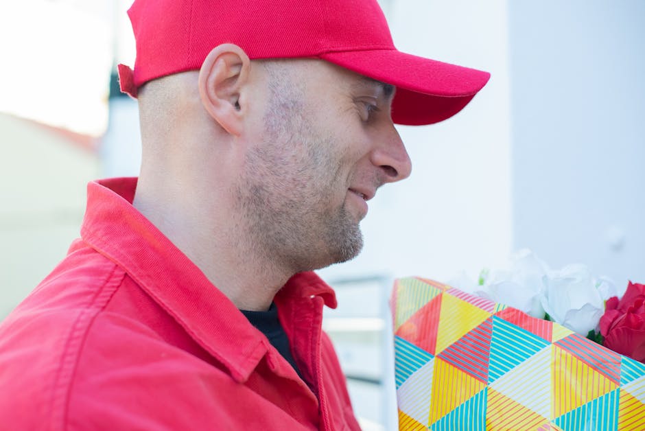 A man dressed in a red cap and matching red jacket is holding a colorful floral gift box with a geometric pattern that includes bright yellow, red, blue, and white triangles. The flowers inside the bo