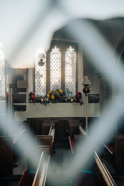The image shows the interior of St Marys Church North Ockendon, featuring a large arched window with intricate leaded glass panels illuminated by natural light. Below the window, a floral arrangement 