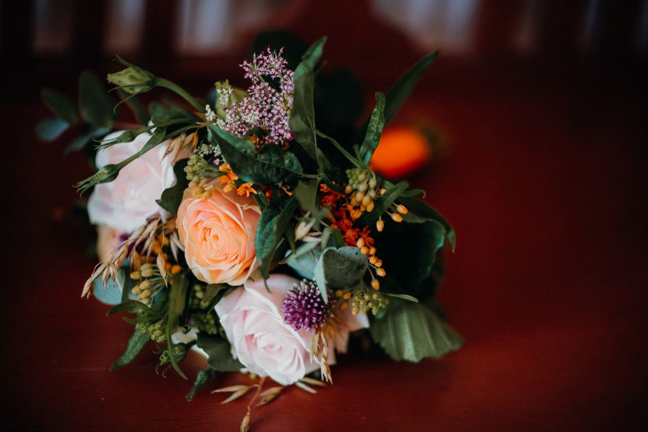 A floral arrangement featuring pale peach and soft pink roses complemented by small purple and white flowers, with green foliage including large textured leaves and delicate sprigs, arranged in a loos
