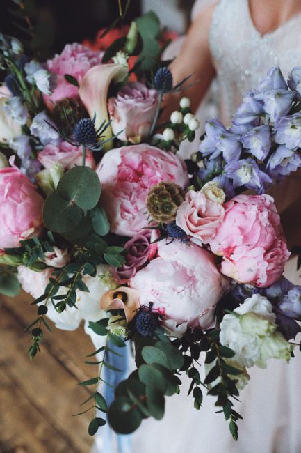 A close-up of a floral arrangement showcasing a bouquet of large, soft pink peonies with ruffled petals, complemented by clusters of light purple delphiniums and white roses. The bouquet is accented w