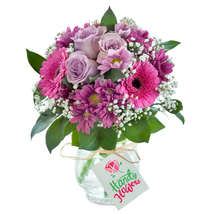 A celebration of fresh artistry and local charm, this bouquet bursts with a harmonious medley of purples and pinks, arranged by hand in North Ockendon with an expert's touch. Leading the arrangement are elegant lavender roses, their petals opening in gentle whorls that seem to shimmer with quiet grace, recalling peaceful country mornings surrounding Courtney Road. Scattered amidst these are striking Gerbera daisies in vivid fuchsia, their dark centres and wide, cheerful petals conjuring images of bustling outdoor markets near North Ockendon village. Small, densely layered magenta chrysanthemums bring textural interest, some with soft green-yellow eyes adding just a hint of wild countryside colour. Sprays of white gypsophila, dainty and ethereal, weave throughout, lightening the ensemble and evoking the hopefulness of new beginnings. Dark green, glossy leaves provide structure and an earthy richness, while the bouquet sits proudly in a clear vase adorned with a rustic, cracked-ice finish-a tactile detail visible to the touch. A simple natural raffia cord encircles the vase's neck, suspending a neat white tag poised for a heartfelt message. This bouquet, expertly arranged by Florist North Ockendon, encapsulates local beauty and the sentimental joy of gifting, making it an ideal choice for any heartfelt occasion or thoughtful delivery throughout the area.