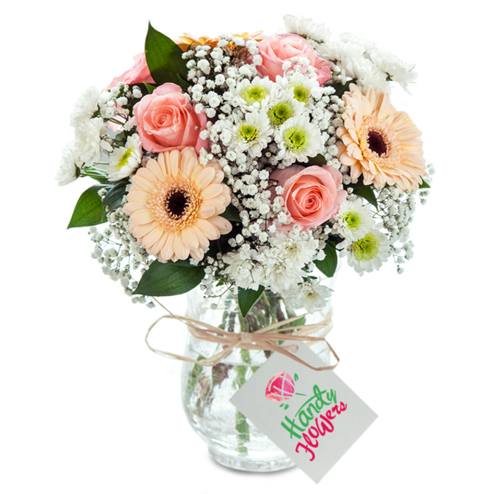 A radiant composition of fresh-picked blooms stands against a pristine white backdrop, thoughtfully arranged by Florist North Ockendon to reflect the gentle spirit of its namesake village. At the bouquet's center, lush, peach-toned roses exude soft romance, reminiscent of garden moments spent along Ockendon Road on sunny afternoons. Alongside, two large, smiling gerbera daisies in soft apricot shades capture the positive energy that defines North Ockendon's friendly neighborhood gatherings. Fine clouds of white baby's breath fill the spaces with delicate texture, much like the mist gathering over the open meadows at dawn, while petite white chrysanthemums adorned with vibrant green centers lend a fresh, lively contrast and extra volume. Rich greenery-just the right amount, sourced from trusted local suppliers-adds depth and dimension. Cradled in a clear glass vase with an understated but rustic finish, the bouquet's stems and water are visible, highlighting quality and careful arrangement. Completing the display, a loop of raffia ribbon or natural twine folds neatly at the vase's neck, holding a white gift tag, consciously left blank for a personal inscription fit for any special event. Expertly constructed, this bouquet is equally suited for anniversaries and everyday moments, reflecting North Ockendon's balance of tradition, warmth, and natural beauty.